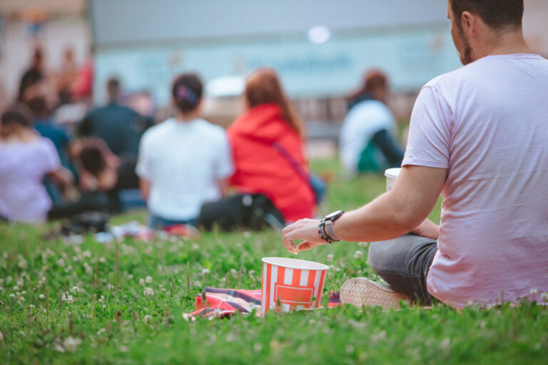 man sitting on the blanket eating snacks in front of big screen on open air cinema