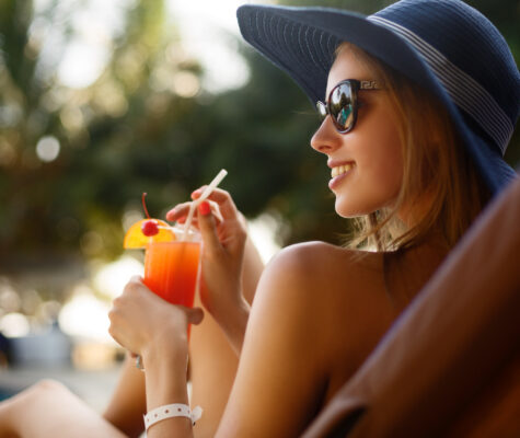 Portrait of young woman with cocktail glass chilling in the tropical sun near swimming pool on a deck chair with palm trees behind. Vacation concept